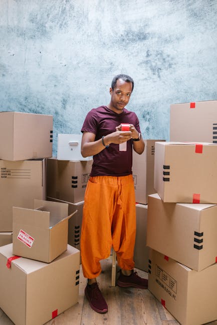 A person standing indoors among a large number of brown cardboard boxes of various sizes, some sealed with red tape and others open revealing packing materials, in a room with a plain, textured light blue wall. The individual is wearing a maroon T-shirt and bright orange loose-fitting trousers, holding a smartphone and looking at the camera. The boxes are stacked on the floor and are positioned around the person, suggesting the process of packing or organizing for a home relocation. The scene is well-lit, indicating an indoor environment used for packing or storage in connection with furniture transport and moving logistics, as typical of professional removals services offered by companies like Hounslow Movers.