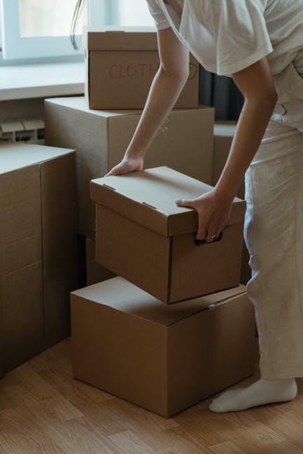 Inside a room during a home relocation, a man with dark skin wearing a grey hoodie is seen leaning forward, smiling, while a woman with light skin and dark hair, dressed in a white top, holds a book a