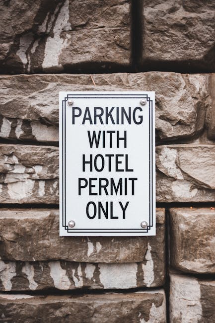 A rectangular, white metal sign with black text reading 'PARKING WITH HOTEL PERMIT ONLY' mounted on a rough brown stone wall composed of irregularly shaped stones. The environment suggests an exterior setting, likely near the entrance of a building where house removals and furniture transport operations could take place. The sign indicates parking restrictions related to hotel permits, relevant to relocating homes in the Hounslow area, as discussed on the Hounslow Movers website. The image emphasizes the importance of parking regulations during packing and moving processes, with the sign serving as a visual cue for permit-required areas familiar in home relocation logistics.