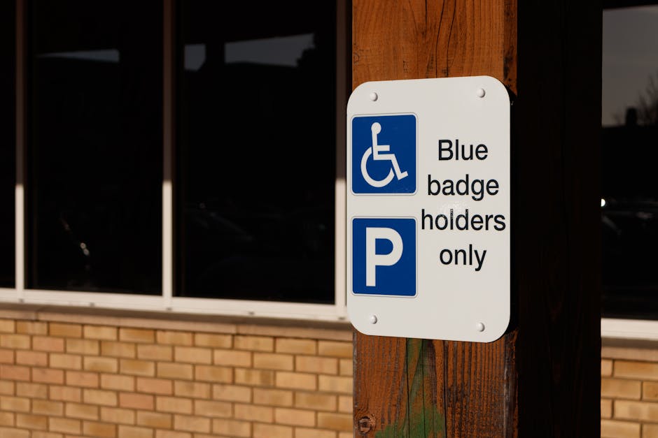 A rectangular white parking sign mounted on a wooden pole outdoors, displaying a blue wheelchair symbol alongside the text 'Blue badge holders only' and a blue parking 'P' icon. The sign is positioned near the corner of a building with a brick wall and large, tinted glass windows. The scene appears to be taken during daylight, with natural lighting illuminating the sign and surroundings. This signage indicates parking restrictions for disabled badge holders, relevant in the context of house removals and moving services such as those provided by Hounslow Movers, who manage safe and compliant furniture transport and home relocation logistics.