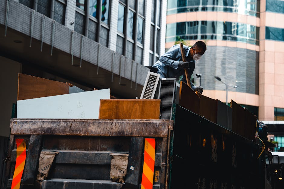 A man wearing a face mask and casual clothing is standing inside the back of a large black waste disposal truck, loading various household furniture and items. The items include wooden cabinets, white