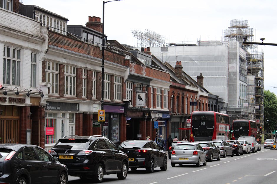 A busy street scene in Hounslow with a row of brick-built terraced shops and residential buildings, some under renovation with scaffolding and white protective sheeting covering the upper floors. The street is lined with parked cars and several black and red double-decker buses, with a small group of pedestrians walking along the sidewalk. Visible street signs indicate 20 mph speed limits and parking regulations. The environment appears to be daytime with natural light illuminating the scene. In the foreground, vehicles are parked parallel to the curb, and the road features lanes designated for traffic flow. The presence of construction scaffolding suggests ongoing building or infrastructure work related to property upgrades or maintenance, reflecting urban development activity in the area. As part of a house removal process, Hounslow Movers might be involved in transporting household furniture or moving services nearby, especially considering the bustling cityscape and residential nature of the street.