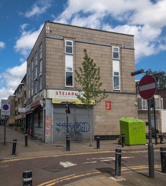 Exterior view of a three-story building on Hounslow High Street during daytime, with a partly cloudy sky overhead. The building features a beige brick façade with several windows, some with white frames, and a ground-floor commercial space with a metal roller shutter door covered in graffiti. Above the shutter is a sign for 'STEJARU', indicating a business that is currently closed, with additional small posters and notices on the shutters. In front of the building, a narrow sidewalk hosts a young, green-leaved tree and various street furniture, including black bollards, a green trash or recycling bin with graffiti, and a lamp post. A black bicycle is partially visible, parked on the right side beneath a signpost with a red and white no-entry sign. The scene suggests a typical urban street scenario, with potential relevance to home relocation or furniture transport activities by Hounslow Movers, situated to support house removals and moving services.