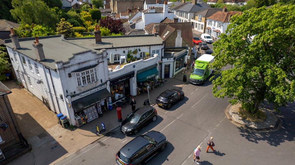 A busy street scene in Hounslow with a row of brick-built terraced shops and residential buildings, some under renovation with scaffolding and white protective sheeting covering the upper floors. The street is lined with parked cars and several black and red double-decker buses, with a small group of pedestrians walking along the sidewalk. Visible street signs indicate 20 mph speed limits and parking regulations. The environment appears to be daytime with natural light illuminating the scene. In the foreground, vehicles are parked parallel to the curb, and the road features lanes designated for traffic flow. The presence of construction scaffolding suggests ongoing building or infrastructure work related to property upgrades or maintenance, reflecting urban development activity in the area. As part of a house removal process, Hounslow Movers might be involved in transporting household furniture or moving services nearby, especially considering the bustling cityscape and residential nature of the street.