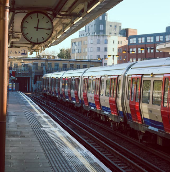 View of Hounslow West station platform with a modern commuter train parked alongside the tracks, featuring a red, white, and blue exterior. The station roof overhead incorporates a large round clock showing approximately 3:00, and fluorescent lighting. The platform surface consists of tiled paving with tactile paving strips near the edge for safety. In the background, multi-storey residential and commercial buildings are visible, with clear daylight and minimal shadows indicating a sunny day. Signs indicating train services and accessibility are present near the station entrance. The scene captures the typical environment of a busy urban train station involved in home relocation and furniture transport activities, highlighting the station's role in facilitating moving logistics in the Hounslow area, as managed by Hounslow Movers for efficient removals and collections.