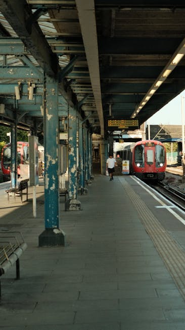 Photograph of Hounslow West station platform showing an outdoor area with a covered canopy supported by blue metal beams, some of which are chipped or weathered. The platform has tactile paving strips along the edge for accessibility, and a row of metal benches on the left side. A person dressed in a light top and dark bottoms is walking along the platform, approaching a red train that is stationary on the tracks. The train has multiple passenger windows and doors for boarding. In the background, additional train carriages and railway infrastructure are visible under a partly cloudy sky, with sunlight illuminating the scene. The overall scene depicts a typical urban train station environment, relevant to the context of house removals or moving logistics related to station-to-station transportation, as seen on the Hounslow Movers website.