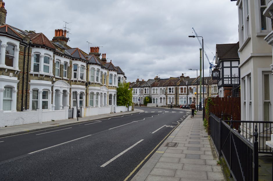 A view of Lampton Road in Hounslow showing residential terraced houses with bay windows and brick facades on the left side and a sidewalk with a black metal fence on the right. The street is paved with asphalt and marked with white dashed and solid lines, indicating parking spaces and traffic lanes. Several parked cars are visible in the distance along the curb on the right, and a few pedestrians are walking along the pavement. Overhead, there are grey, overcast skies and a crane in the background, suggesting ongoing construction nearby. The scene captures a typical urban residential area suitable for house removals and furniture transport services, with the environment prepared for the loading process that Hounslow Movers might facilitate during home relocation projects. The street's layout and the presence of loading restrictions are subtly referenced through the scene's design, emphasizing the importance of understanding local parking regulations for efficient moving logistics.