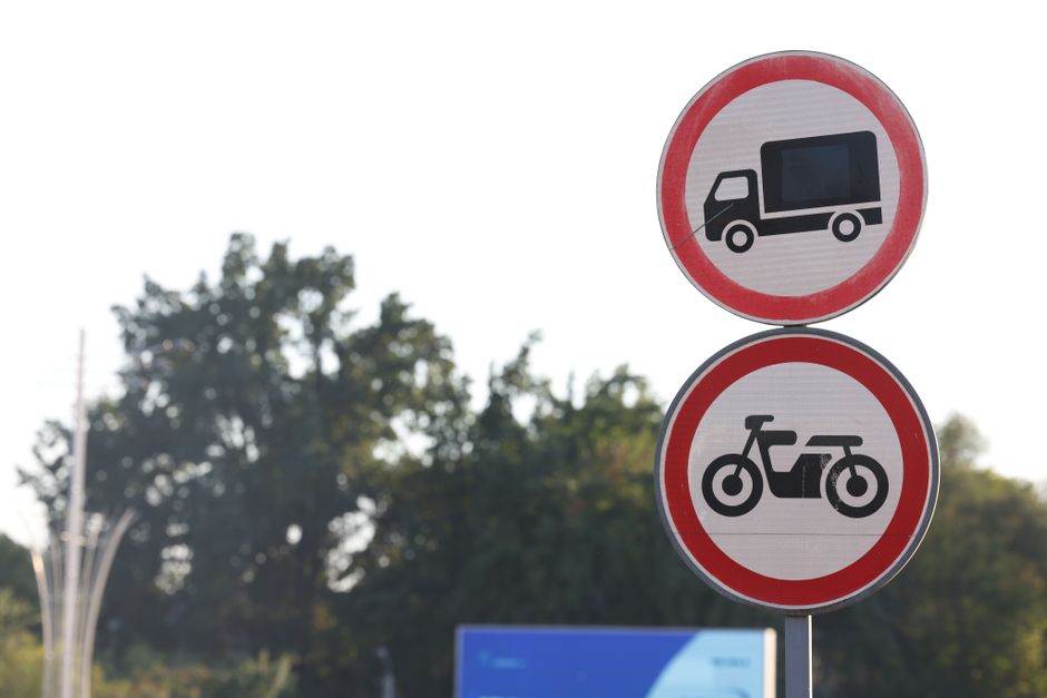 Two circular road signs mounted on a single pole against a backdrop of green trees and a partly cloudy sky. The upper sign has a black truck icon within a red circle indicating no trucks allowed, while the lower sign displays a black motorcycle icon within a red circle indicating no motorcycles permitted. The signs are part of local road restrictions in Hounslow, relevant to house removals and furniture transport from Lampton Road, as managed by Hounslow Movers, a professional removals company. The signs are situated near a pavement area, and the image is taken during daylight with natural lighting, providing clear visibility of the restricted vehicle types that may impact loading and unloading processes during home relocation services.