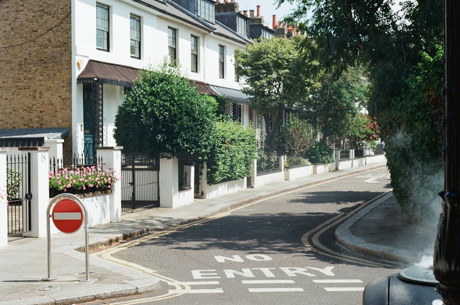 A view of Lampton Road in Hounslow showing residential terraced houses with bay windows and brick facades on the left side and a sidewalk with a black metal fence on the right. The street is paved with asphalt and marked with white dashed and solid lines, indicating parking spaces and traffic lanes. Several parked cars are visible in the distance along the curb on the right, and a few pedestrians are walking along the pavement. Overhead, there are grey, overcast skies and a crane in the background, suggesting ongoing construction nearby. The scene captures a typical urban residential area suitable for house removals and furniture transport services, with the environment prepared for the loading process that Hounslow Movers might facilitate during home relocation projects. The street's layout and the presence of loading restrictions are subtly referenced through the scene's design, emphasizing the importance of understanding local parking regulations for efficient moving logistics.