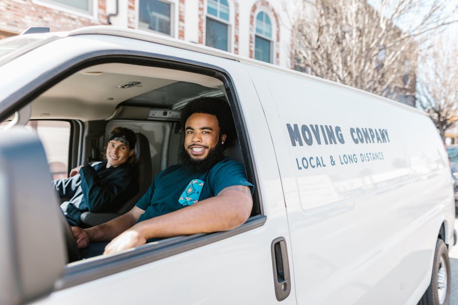 Inside a white moving van parked outdoors in an urban area with multi-storey buildings in the background, two men are visible through the open side door, engaging in home relocation activities. The man in the driver’s seat, with dark skin and a full beard, is smiling and wearing a dark blue polo shirt. The passenger seated beside him, a lighter-skinned woman with short brown hair and wearing a dark jacket, is also smiling. Inside the van, various packing materials such as cardboard boxes, plastic-wrapped furniture, and blankets are visible, indicating an ongoing furniture transport and packing process for a house removal. The cargo area includes cardboard boxes stacked on top of each other, some wrapped in plastic for protection, along with a trolley and moving straps used for securing belongings during loading. The environment is well-lit with natural daylight, suggesting a clear day, and the van appears to be in the midst of loading or preparing for a move. Hounslow Movers' branding is visible on the van's side, indicating their local and long-distance removals services, which encompass packing, loading, and transportation for home relocations.
