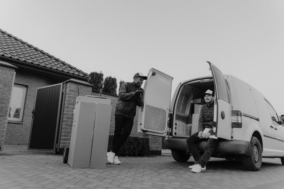 Two men engaged in house removal activities outside a residential property, depicted in black and white. One man, standing next to a large cardboard box and dressed in casual clothing, appears to be preparing or inspecting packing materials. The other man, sitting on the open rear door of a white moving van, wears a hat and jacket, possibly overseeing or coordinating the loading process. The van, parked on a paved driveway, is partially filled with furniture, cardboard boxes, and packing materials, including blankets and plastic wrap, indicating a furniture transport phase of the home relocation. The background features a brick house with a tiled roof, a small garden area, and a door leading inside. The scene captures the logistics of packing and loading, with equipment such as trolleys and straps likely used to facilitate safe and efficient transfer, reflecting professional removals by Hounslow Movers while emphasizing key steps in house moves and furniture transport.