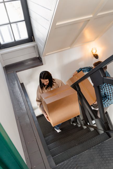 A woman wearing a beige sweater is carrying a large cardboard box up black-painted stairs inside a residential property during a home relocation. She is positioned near the bottom of the staircase, holding the box with both hands. Behind her, a man in a blue and black checkered shirt is also lifting a cardboard box, assisting with the moving process. The staircase has a black handrail and dark carpeting on the steps. To the left, a window with multiple panes allows natural light into the interior, illuminating the white walls and ceiling. A small wall-mounted light fixture on the right adds additional lighting. The scene depicts the house removal team coordinating the furniture transport within the property, emphasizing careful packing and stair navigation for narrow access areas, as reflected on the page titled 'TW3 flat removals tips for narrow stairs and access' on hounslowmovers.co.uk.
